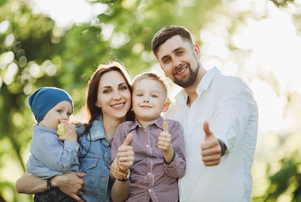 Positive and stylish family walking in park together with kids, posing and gesturing super by fingers at camera. Mother and father embracing their two son. Concept of family look.